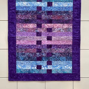 May include: A quilted wall hanging with a vibrant color palette of blues, purples, and lavender. The design features horizontal strips of patterned fabric, accented with dark purple squares. A dark purple border frames the artwork.