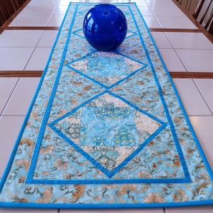 May include: A blue and turquoise quilted table runner with a floral pattern and diamond accents. A large blue glass sphere sits on the runner, which is placed on a white tiled table. The runner has a bright blue border.