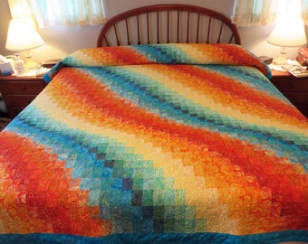 May include: A colourful quilt with a diagonal pattern of squares in shades of blue, green, yellow, orange, and red. The quilt is made with a batik fabric and has a turquoise border.