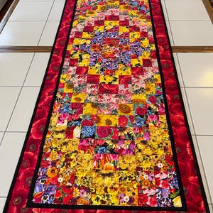 May include: A rectangular floral table runner with a patchwork design. The center features a vibrant mix of colorful flowers in a grid pattern. The border is a deep red with large poppy flower prints, and a black trim.