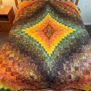 May include: A colorful quilt with a diamond pattern in shades of yellow, orange, green, and brown. The quilt is made of small, square fabric pieces arranged in a geometric design. The quilt is on a bed.