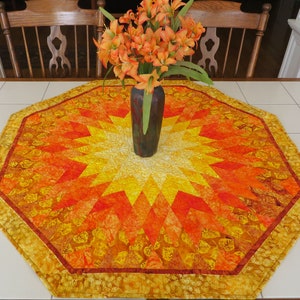 May include: An octagonal table runner with a sunburst design in shades of yellow, orange, and red. The runner has a patterned border and a vase of orange flowers in the center. A wooden chair is visible in the background.