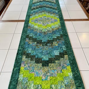 May include: A rectangular quilted table runner featuring a concentric diamond pattern in shades of lime green, teal, and dark forest green. The fabric displays various mottled and swirled textures, finished with a solid dark green border.