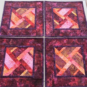 May include: A set of four square fabric placemats featuring a central geometric pattern in shades of pink, orange, and red, surrounded by a dark purple floral-patterned border. Each placemat displays intricate machine quilting with swirling lines.