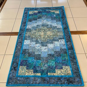 May include: A rectangular table runner featuring a patchwork design of small squares in various shades of blue, teal, and light green. The fabric displays intricate batik-style patterns and is bordered by a solid bright blue fabric trim.