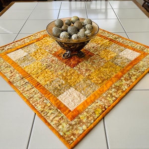 Könnte beinhalten: Eine quadratische, gesteppte Tischdecke mit Patchwork-Design in Orange-, Gelb- und Cremetönen mit komplizierten Wirbelstichmustern. In der Mitte steht eine Glasschale, die mit goldenen und silbernen Dekokugeln gefüllt ist.