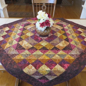 May include: A square table runner with a geometric pattern of triangles in shades of purple, brown, and gold. The runner is made of fabric with a batik-like design. A vase with white and red flowers sits in the center of the runner.