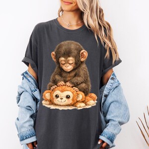 Peut inclure: T-shirt gris fonc&eacute; avec une illustration d'un b&eacute;b&eacute; singe aux yeux ferm&eacute;s, reposant sur un singe en peluche. Le singe en peluche a un visage souriant. Le t-shirt a des manches retrouss&eacute;es.