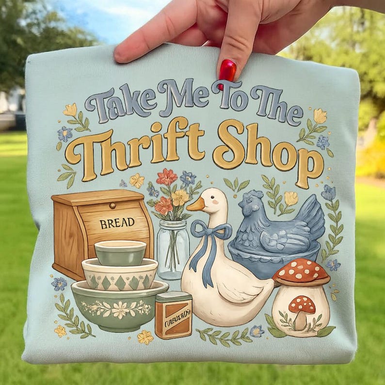 Op de afbeelding: Lichtblauwe draagtas met de tekst "Take Me To The Thrift Shop" in vintage stijl. De tas heeft illustraties van een brooddoos, kommen, een eend, een kip en een paddenstoel.