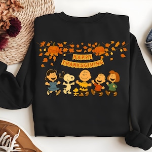 Könnte beinhalten: Schwarzes Sweatshirt mit einer Grafik der Peanuts-Charaktere, darunter Charlie Brown und Snoopy, die Thanksgiving feiern. Das Design umfasst Kürbisse, Herbstblätter und den Text "Happy Thanksgiving" auf einem Banner.
