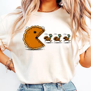 May include: Cream-colored long-sleeve shirt featuring a cartoon design. The graphic depicts a pie slice with a Pac-Man-like design chasing three turkeys. The turkeys are running away from the pie. The design is in shades of brown, orange, and green.