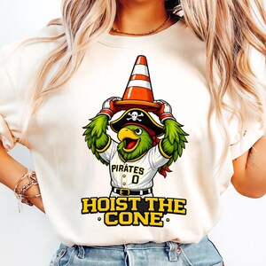 Pode incluir: Camiseta creme com um papagaio de desenho animado usando um chap&eacute;u de pirata e uma camisa de beisebol com a palavra "Pirates". O papagaio est&aacute; segurando um cone de tr&acirc;nsito laranja e branco. O texto "HOIST THE CONE" est&aacute; abaixo.