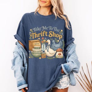 Op de afbeelding: Marineblauw T-shirt met de tekst "Take Me To The Thrift Shop" in geel en groen. De afbeelding bevat een brooddoos, kommen, een eend, een kip en een paddenstoel.