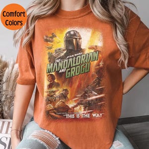 May include: Orange t-shirt featuring a Star Wars graphic with The Mandalorian and Grogu. The design includes characters and the phrase "THIS IS THE WAY". The shirt has a vintage look with "Comfort Colors" text in a circle.