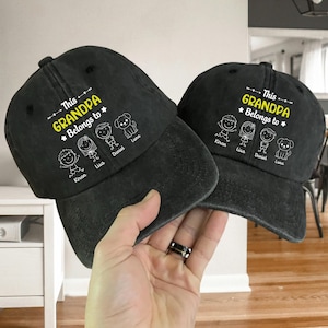 May include: Two black baseball caps with the text "This Grandpa Belongs to" and the names Kiran, Lisa, Daniel, and Luna. The caps have a distressed look and a curved brim. The text and illustrations are in white and yellow.