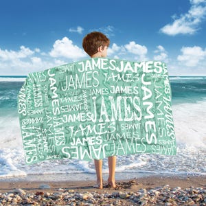 May include: A mint green blanket with the name "JAMES" printed repeatedly in white. The blanket is held by a person on a beach with the ocean and blue sky in the background.