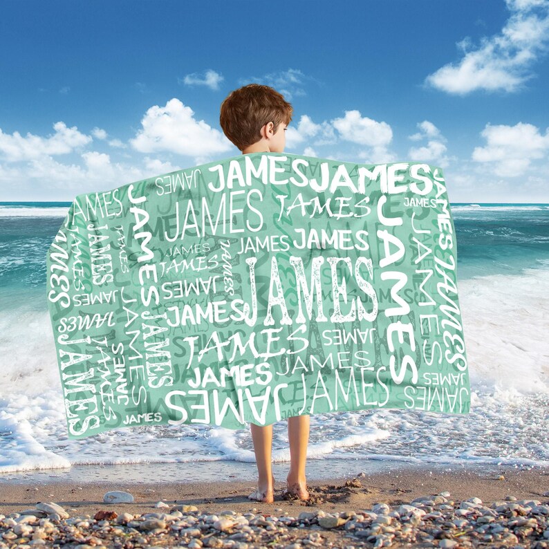 May include: A mint green blanket with the name "JAMES" printed repeatedly in white. The blanket is held by a person on a beach with the ocean and blue sky in the background.