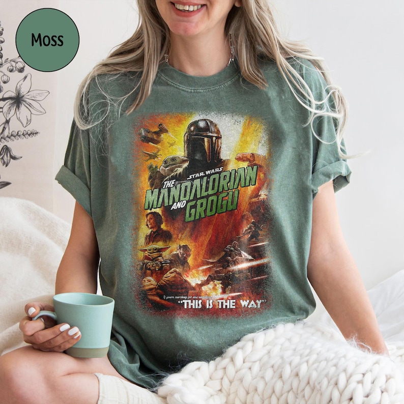 May include: Moss green t-shirt featuring a graphic from "The Mandalorian and Grogu." The design includes the Mandalorian, Grogu, and other characters from the Star Wars series. The text "THIS IS THE WAY" is also visible.