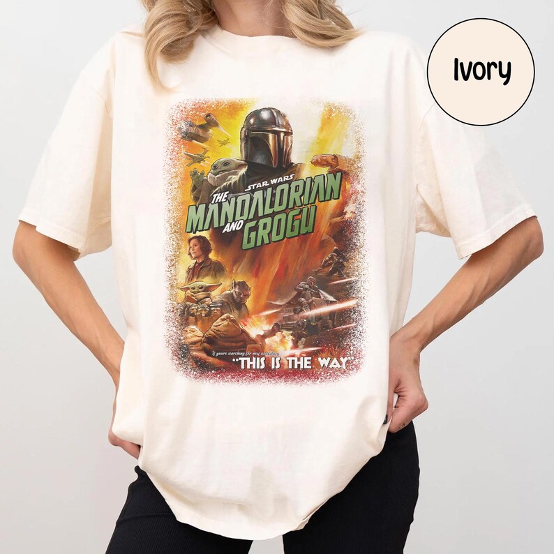 May include: Ivory-colored t-shirt featuring a Star Wars graphic. The design showcases "The Mandalorian and Grogu" with characters and the phrase "THIS IS THE WAY." The shirt has short sleeves and a relaxed fit.