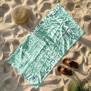 May include: A teal beach towel with the name "JAMES" printed in white, repeated across the surface. The towel is lying on a sandy beach, with a straw hat and brown sandals nearby. A palm frond is visible in the corner.