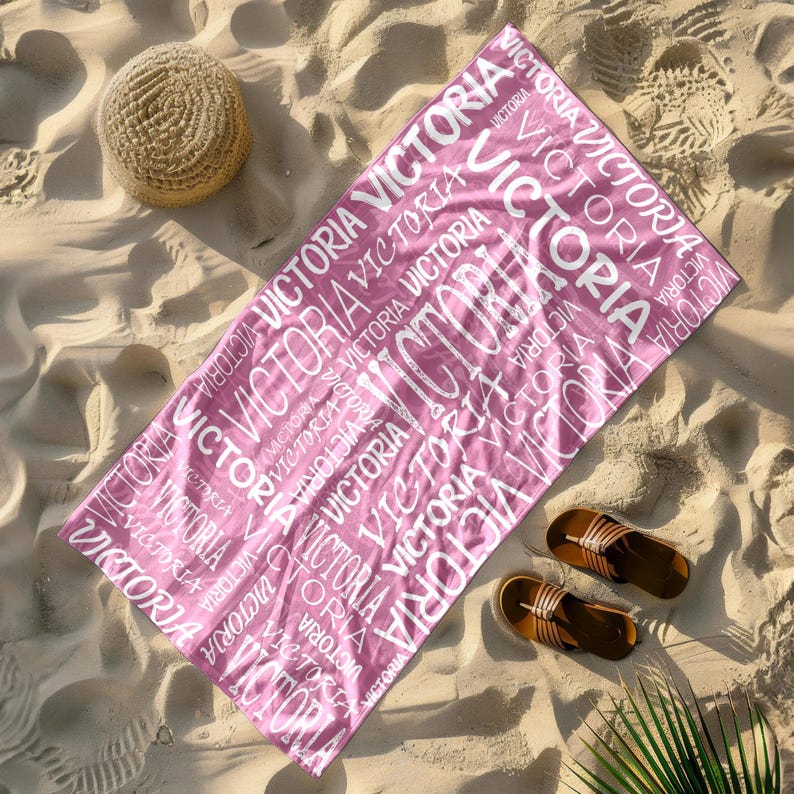May include: A pink beach towel with the name "VICTORIA" printed repeatedly in white. The towel is rectangular and lies on a sandy beach, with a straw hat and sandals nearby. The towel is approximately 70 inches long.