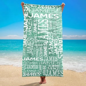 May include: A rectangular beach towel with the name "JAMES" printed repeatedly in white on a teal background. The towel is held up against a backdrop of a sandy beach, ocean, and blue sky.