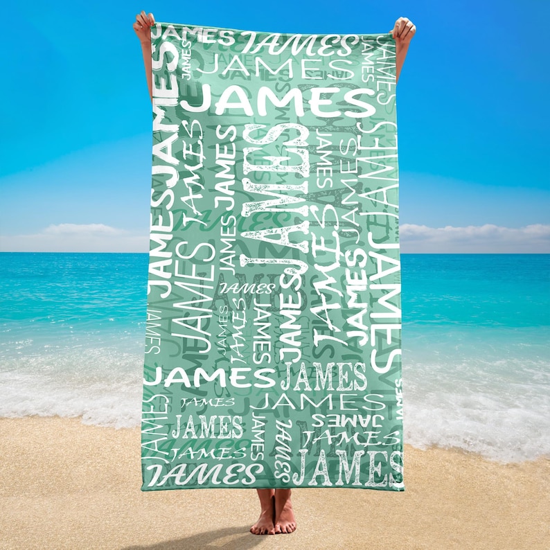 May include: A rectangular beach towel with the name "JAMES" printed repeatedly in white on a teal background. The towel is held up against a backdrop of a sandy beach, ocean, and blue sky.