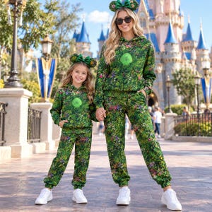 Peut inclure: Ensembles assortis composés de sweats à capuche et de pantalons de jogging verts, ornés d'un imprimé floral et végétal vibrant avec un emblème en spirale central. Chaque tenue est complétée par un serre-tête vert à nœud et fleurs, avec baskets blanches.