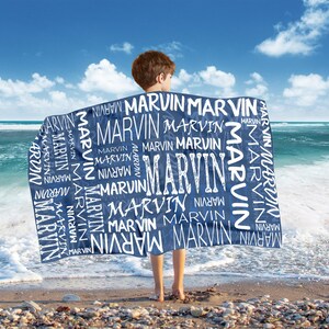 May include: A navy blue beach towel with the name "MARVIN" in white, repeated in various sizes and fonts. The towel is held by a person on a sandy beach with the ocean and blue sky in the background.