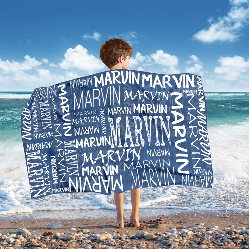 May include: A navy blue beach towel with the name "MARVIN" in white, repeated in various sizes and fonts. The towel is held by a person on a sandy beach with the ocean and blue sky in the background.