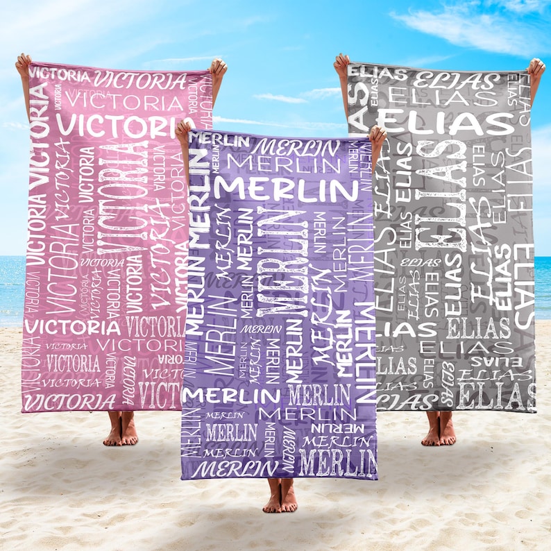 May include: Three personalized beach towels held up on a sandy beach. One is pink with "VICTORIA" in white, another is purple with "MERLIN", and the third is gray with "ELIAS". The towels are rectangular.
