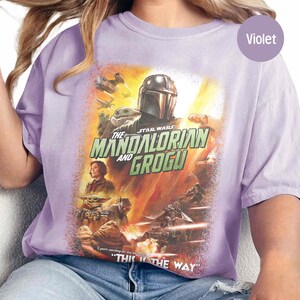 May include: Violet-colored t-shirt featuring a graphic from the Star Wars series, "The Mandalorian and Grogu." The design includes characters and the phrase "This is the Way." The shirt has short sleeves and a crew neck.