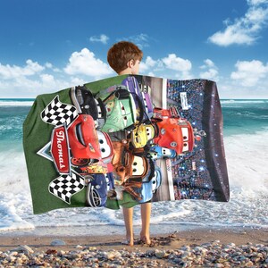 Puede incluir: Una colorida manta con varios coches de carreras de dibujos animados, incluido uno rojo con el nombre "Thomas". La manta tiene un fondo verde con banderas a cuadros y una escena de carreras. Una persona sostiene la manta en una playa.