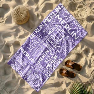 May include: A purple beach towel with the name "MERLIN" printed in white. The towel is rectangular and lies on a sandy beach, next to a straw hat and brown sandals.