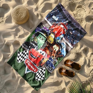 Puede incluir: Una toalla de playa con un dise&ntilde;o colorido de coches de carreras animados sobre un fondo verde. La toalla tiene una bandera a cuadros y el nombre "Thomas". Un sombrero de paja y sandalias est&aacute;n junto a la toalla en la arena.