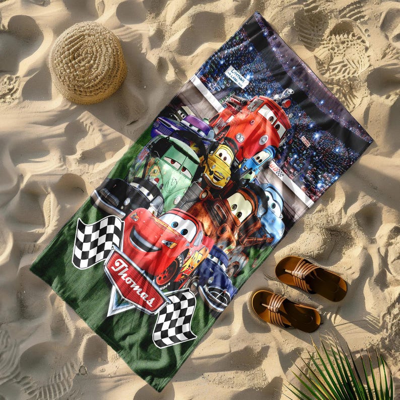 Puede incluir: Una toalla de playa con un dise&ntilde;o colorido de coches de carreras animados sobre un fondo verde. La toalla tiene una bandera a cuadros y el nombre "Thomas". Un sombrero de paja y sandalias est&aacute;n junto a la toalla en la arena.