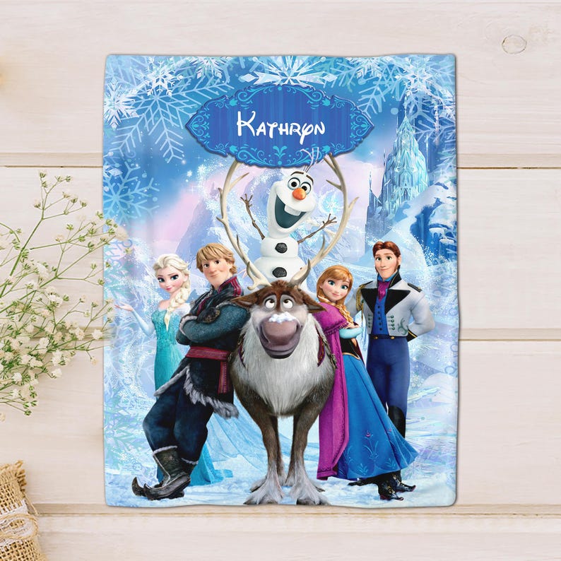 Puede incluir: Una manta rectangular con personajes de la pel&iacute;cula Frozen, incluyendo a Elsa, Anna, Kristoff y Olaf. La manta tiene un fondo azul y blanco con copos de nieve y el nombre "Kathron" en una pancarta decorativa.