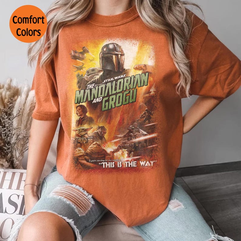 May include: Orange t-shirt featuring a Star Wars graphic with The Mandalorian and Grogu. The design includes characters and the phrase "THIS IS THE WAY". The shirt has a vintage look with "Comfort Colors" text in a circle.