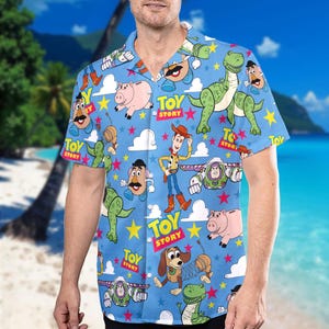 May include: A short-sleeved, button-down shirt with a light blue background featuring various Toy Story characters. The shirt has a repeating pattern of Woody, Buzz Lightyear, Mr. Potato Head, Rex, Hamm, and Slinky Dog, along with the words "Toy Story".