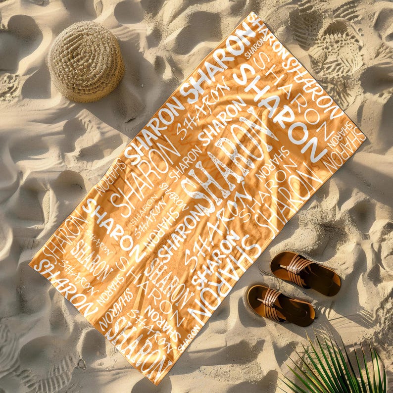 May include: An orange beach towel with the name "SHARON" repeated in white. The towel is on a sandy beach, with a straw hat and brown sandals nearby. A palm frond is visible.
