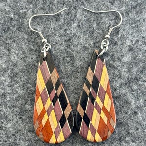 May include: A pair of teardrop-shaped earrings with a geometric pattern. The earrings feature a mosaic design of various wood tones, including orange, yellow, black, and purple. The earrings have silver-colored fish hook ear wires.