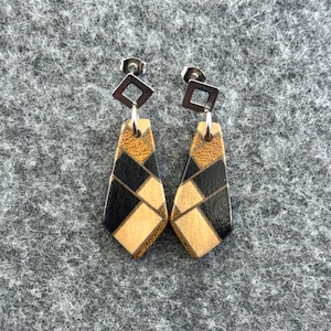 May include: A pair of geometric wooden earrings. Each earring has a diamond-shaped silver stud, a small silver square, and a wooden pendant with black, tan, and gold geometric shapes. The earrings are designed for pierced ears.