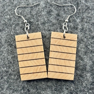May include: A pair of wooden earrings with a rectangular shape. Each earring features a light brown wood grain with six horizontal black lines. The earrings have silver-colored fish hook ear wires.