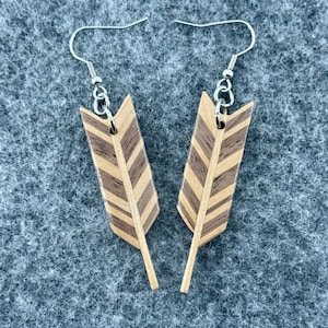 May include: A pair of wooden feather earrings. Each earring has a light brown feather design with dark brown chevron stripes. The earrings have silver-coloured ear wires and are displayed on a grey surface.