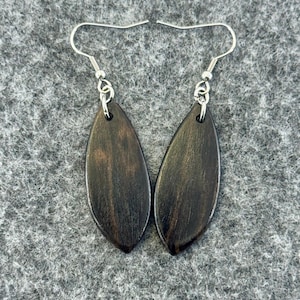 Puede incluir: Un par de pendientes en forma de lágrima con un acabado de veta de madera marrón oscuro. Los pendientes tienen ganchos para las orejas plateados y se muestran sobre una superficie gris texturizada. Un accesorio sencillo y elegante.