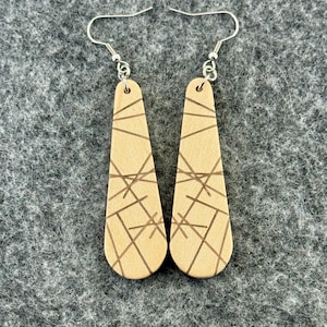 May include: A pair of beige leather earrings with a geometric design. The earrings are teardrop-shaped and feature a series of intersecting lines etched into the surface. The earrings hang from silver-colored fish hook ear wires.