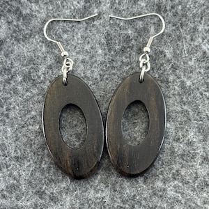 May include: A pair of dark brown oval wooden earrings with a central oval cutout. The earrings hang from silver-colored fish hook ear wires. The wood grain is visible on the surface of the earrings.