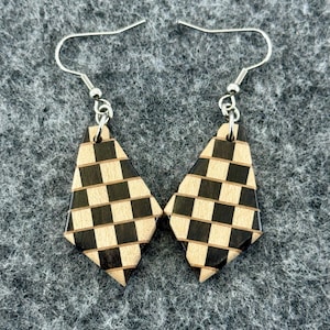 May include: A pair of wooden earrings with a geometric checkerboard pattern in black and natural wood tones. The earrings are diamond-shaped with silver-colored ear wires, displayed on a gray textured surface.