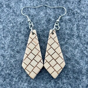 May include: A pair of light-colored wooden earrings with a geometric pattern. The earrings are diamond-shaped with a grid-like design etched into the wood. They hang from silver-colored fish hook ear wires.
