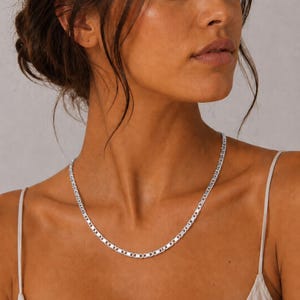 May include: A silver-colored necklace with a flat, rectangular link design. The necklace is worn around the neck and rests on the chest. The model is wearing a white spaghetti strap top.